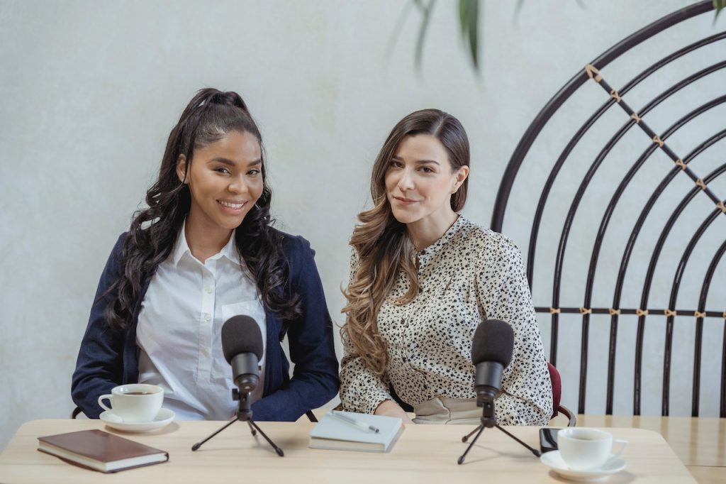 Image of two woman with a notebook and two microphonewith a stand on top of a table. Source: george milton, pexels