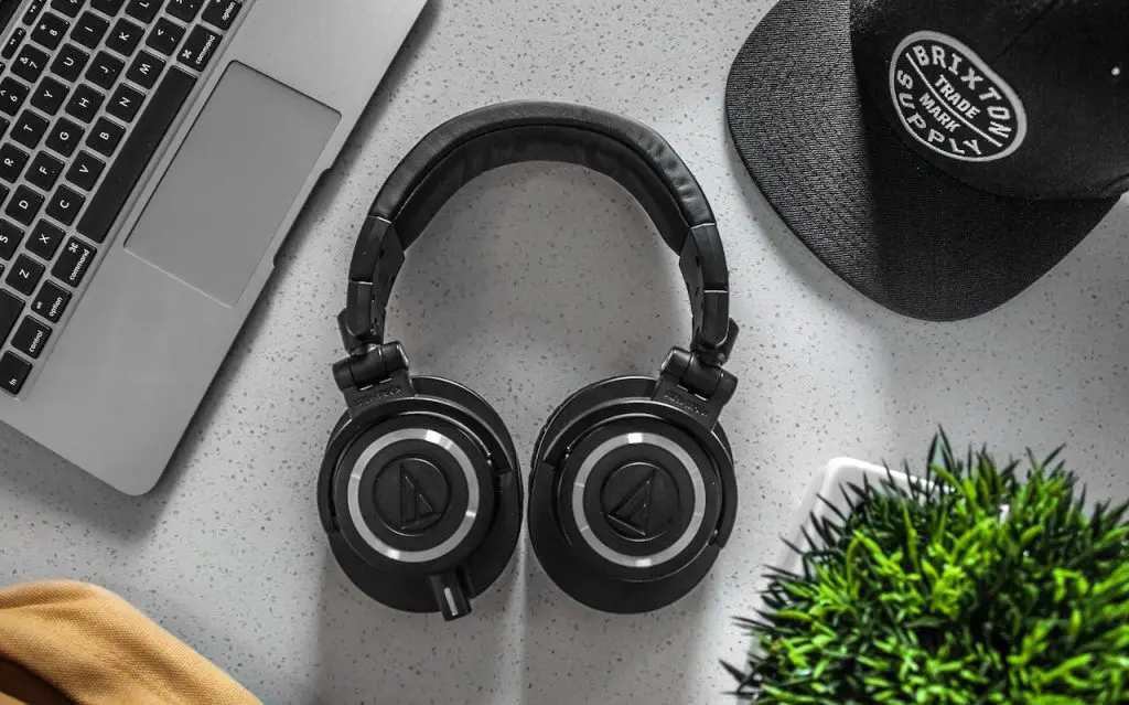 Image of a black colored wireless headphone with a laptop, a cap, and a plant beside it. Source garrett morrow, pexels