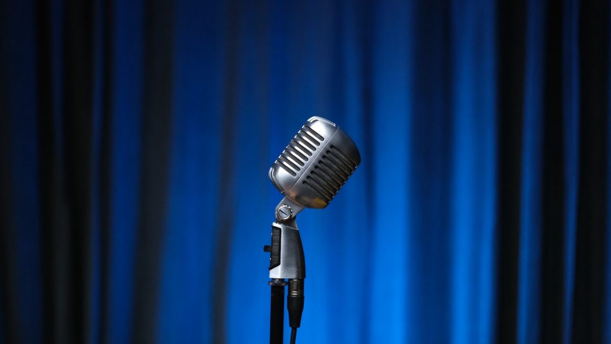 Image of a silver microphone on a stand, with blue curtains at the back. Source: ahmet bozku, pexels
