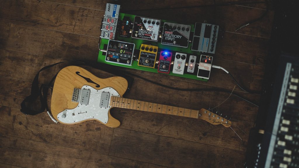 Image of a guitar and a compressor pedal laid on a wooden floor. Source: unsplash