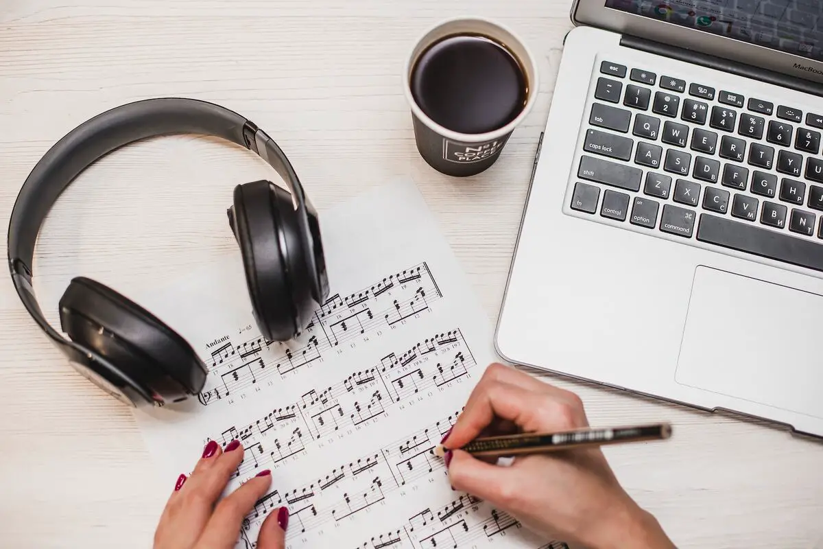 Image of a person writing on a music sheet with headphones and a laptop on the table. Source: unsplash