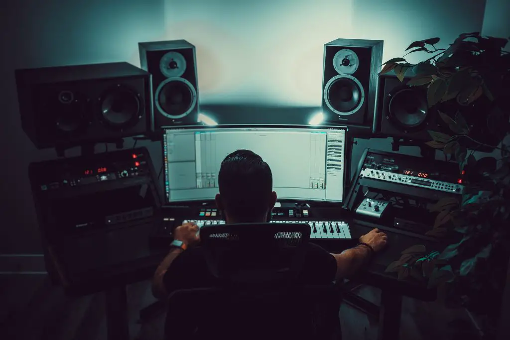 Image of man sitting in a recording studio setup with a computer monitor, audio mixer, and studio monitors. Source: unsplash