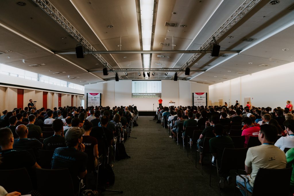 An event hall with a man talking in front of an audience. Source: unsplash